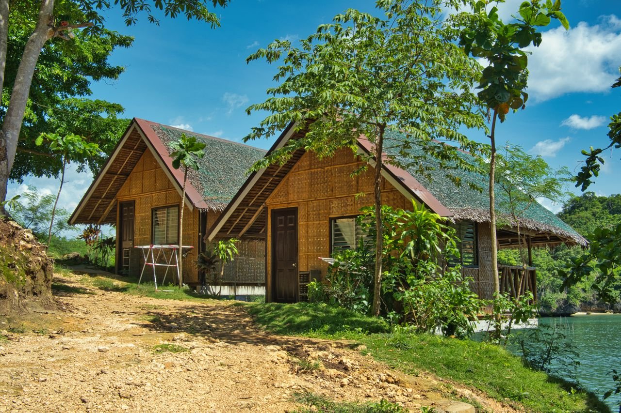 Home Serene bamboo cottages by the lake, surrounded by lush green trees and a clear blue sky.